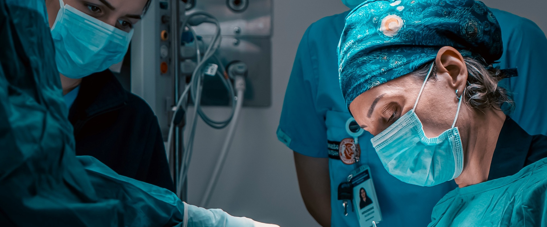 Surgeons in teal scrubs and surgical masks performing an operation in an operating room, with medical equipment visible in the background