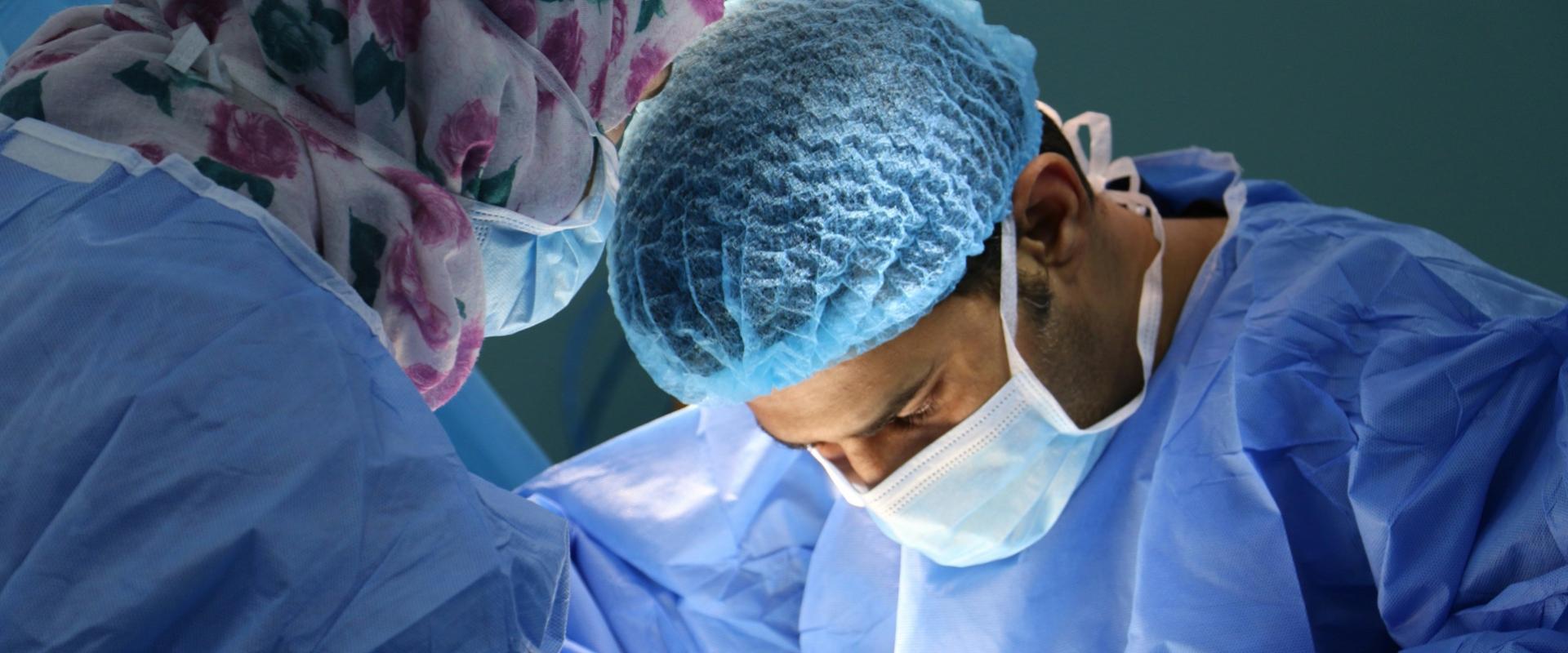 Two surgeons in blue surgical scrubs, surgical masks, and hair caps working closely together during a medical procedure, with one wearing a floral-patterned surgical cap